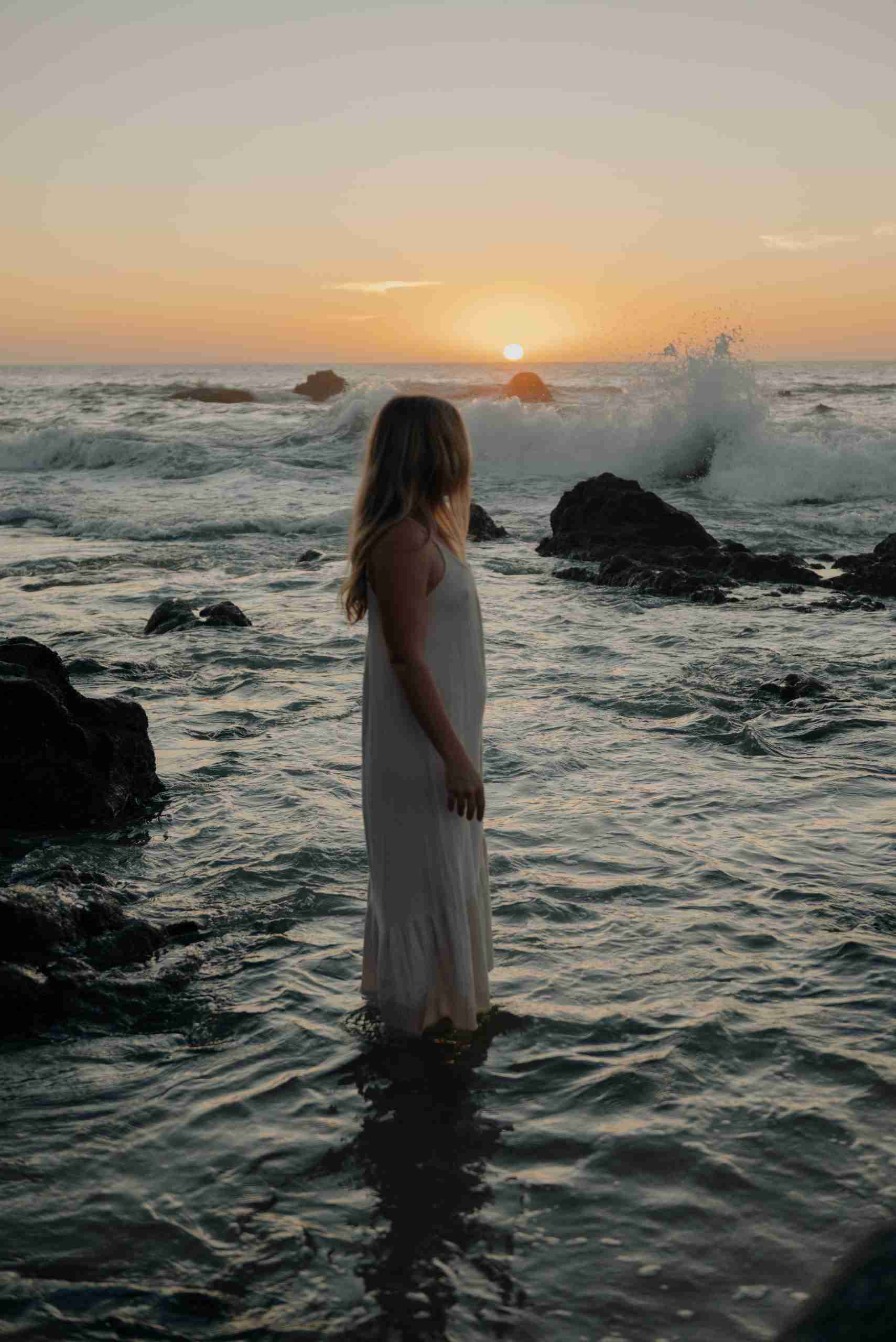 A woman standing still in the ocean at golden hour, facing the horizon