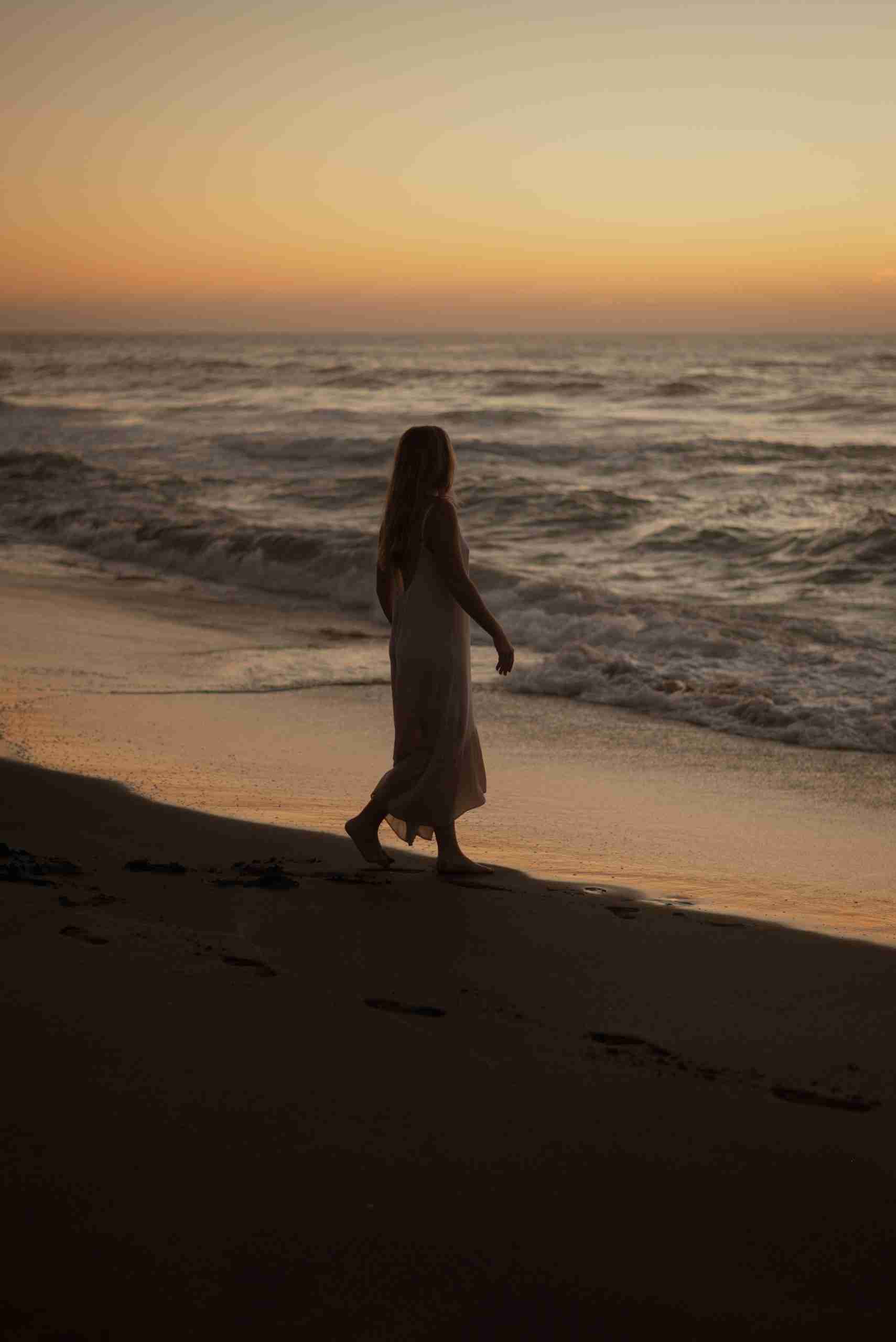 A woman walking along the beach at dusk toward a warm, hopeful sky